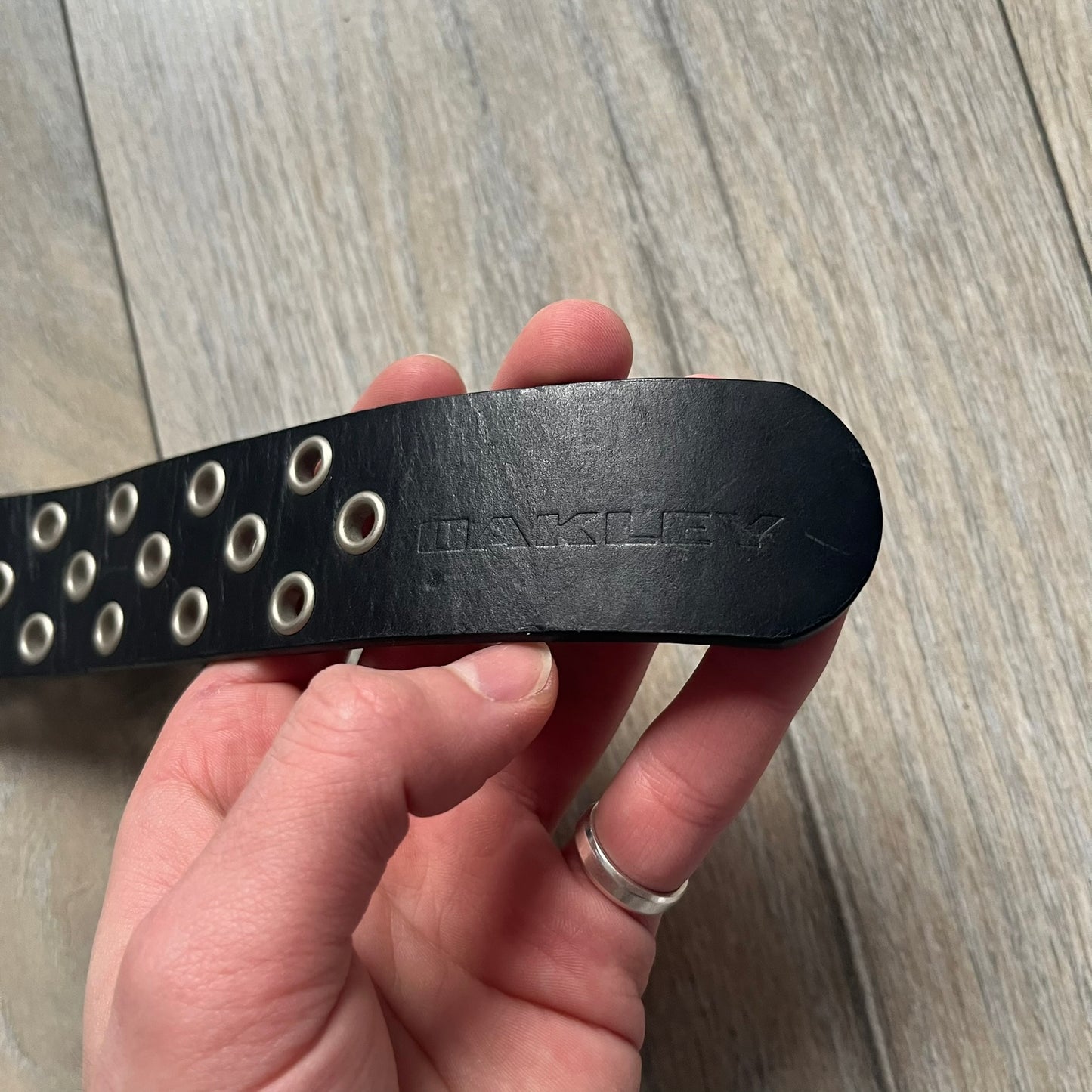 Oakley 2004 Black Leather Skull Belt is a genuine black leather belt featuring full-length holes for adjustable sizing. The belt end is embossed with a carved Oakley logo, and it includes a metal skull buckle with Oakley branding. Made in 2004, it combines durable leather construction with signature Oakley design details.