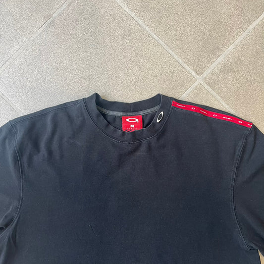 Oakley 2000's Metal Ellipse Black Tee is a black T-shirt made from 95% cotton and 5% spandex, featuring a raised collar with a metal Oakley Ellipse logo and a red stripe printed with Oakley logos on one shoulder.
