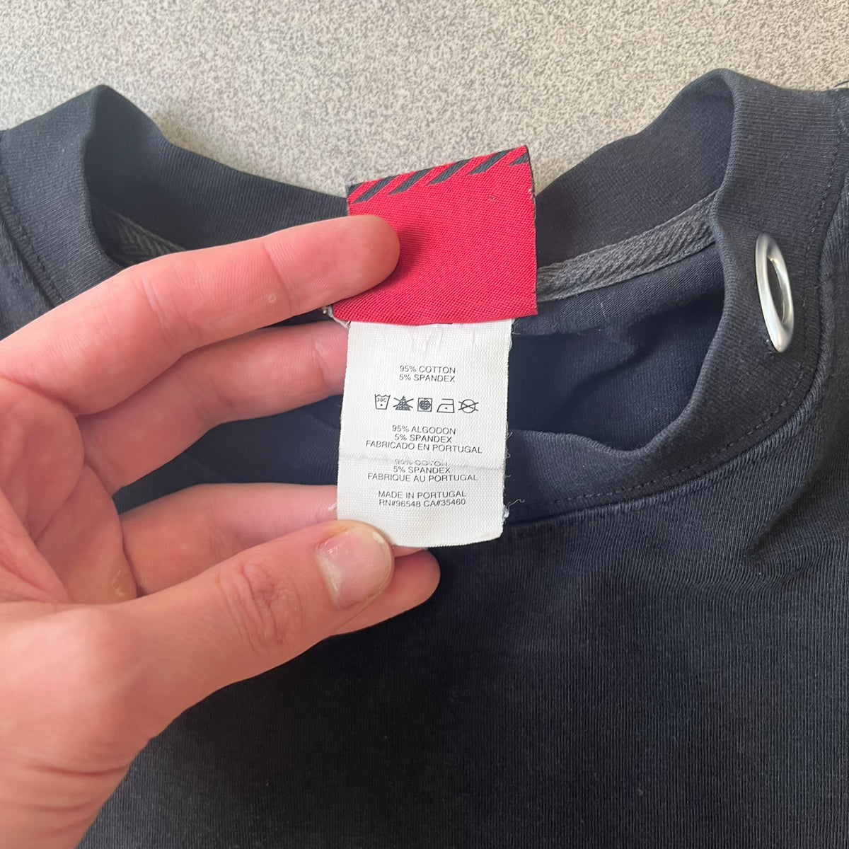 Oakley 2000's Metal Ellipse Black Tee is a black T-shirt made from 95% cotton and 5% spandex, featuring a raised collar with a metal Oakley Ellipse logo and a red stripe printed with Oakley logos on one shoulder.