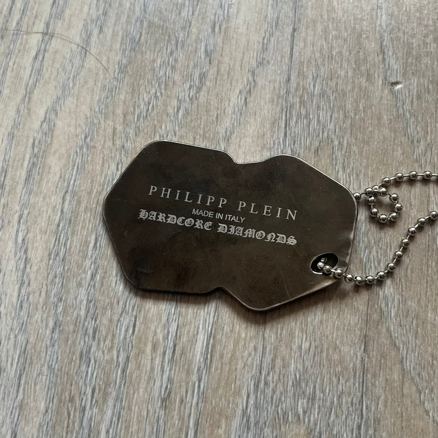 Philipp Plein Two Dices Necklace features a flat metal pendant from Philipp Plein paired with a non-original ball chain. The pendant is designed as two connected dice in a dark grey finish with black enamel dots, with one side replacing the dots with the Philipp Plein skull motif, while the reverse is engraved with the brand logo. Originally used as a zipper pull or accessory attachment, the piece is made in Italy and comes from the 2013 “Hardcore Diamonds” collection.