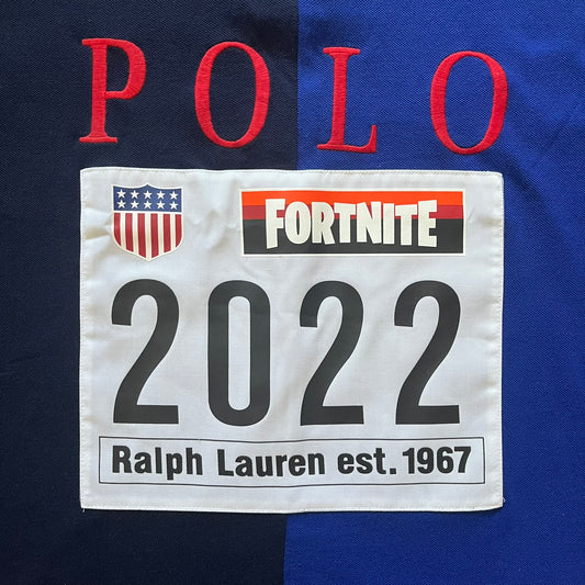 Polo Ralph Lauren x Fortnite 2022 Polo Shirt featuring a two-tone split design in blue and navy with a crisp white collar. The front showcases a large white panel with “2022” text, Ralph Lauren Est. 1967 branding, and a Fortnite logo, along with an oversized embroidered Polo logo above. A limited collaboration piece combining Ralph Lauren’s heritage aesthetic with Fortnite’s playful edge.