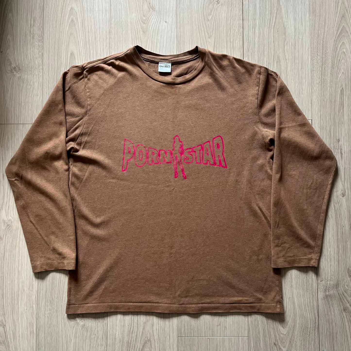Vintage 90’s Porn Star Clothing longsleeve in brown, featuring a bold pink logo on the chest with a silhouette outline of a woman between the words “Porn” and “Star.” Crafted with quality construction, it carries the brand’s signature edgy aesthetic. Finished with a playful “must be 18 or over” detail on the tag, this rare piece embodies 90’s streetwear provocative style.