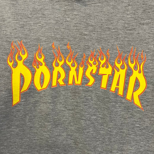 Porn Star Clothing Thrasher Hoodie from the 1990s, made in a gray cotton fabric. The front features a large flame-style “Pornstar” logo that is a spin-off of the Trasher one, with matching flame graphics running down both sleeves. Designed with an adjustable drawstring hood, front kangaroo pocket, and ribbed cuffs and hem. Finished with the original “Must Be 18 Or Over” Porn Star Clothing tag.