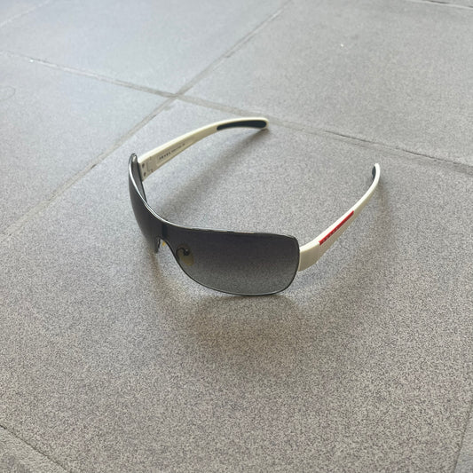 Prada Linea Rossa SPS57G White Sunglasses are shield-style sunglasses featuring a single dark wraparound lens and white arms. One temple is finished with the signature red Prada Linea Rossa logo insert, while the nose pads feature subtle Prada branding. Made in Italy, the sunglasses are lightweight sporty design and they come complete with the original Prada case.
