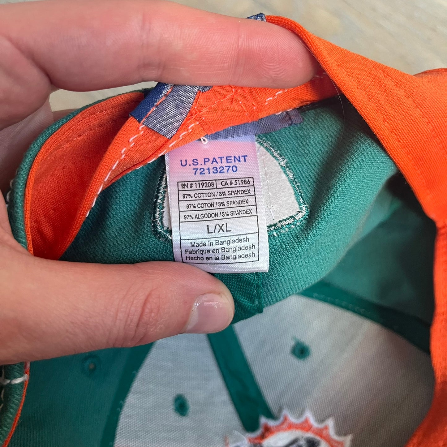 Reebok NFL Miami Dolphins Onfield Snapback Cap is a turquoise snapback featuring embroidered Miami Dolphins branding on the front, framed by two bold orange stripes positioned at the top and bottom of the front panel. The visor includes white stitching on the top surface and an orange underside for contrast. The side is detailed with a white Reebok logo, while the back features NFL Equipment and Onfield branding.