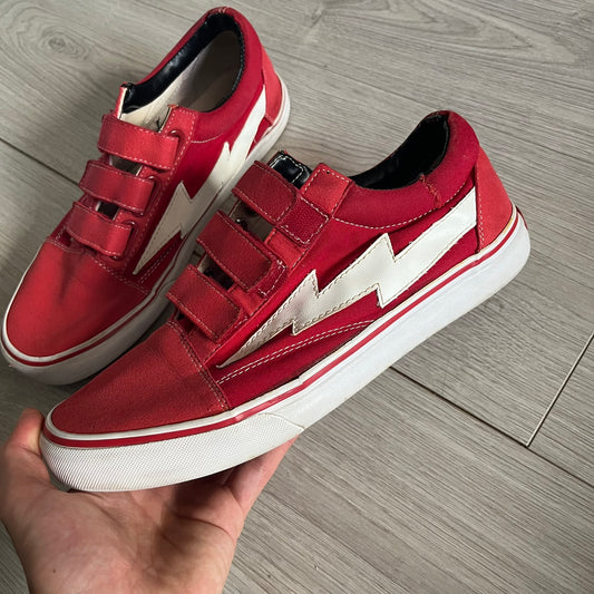 Revenge x Storm Red Strap Sneakers designed by Ian Connor. Featuring a red upper with white leather lightning bolts replacing the traditional Vans stripe. Finished with Velcro strap closures, white midsoles, black outsoles, and the signature “Revenge x Storm” branding in red on the sole.