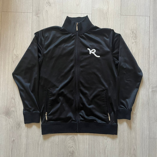 Vintage Rocawear black track jacket featuring a small embroidered chest logo and big white branding across the back. Silver zipper and wrist emblem add refined details to this early-2000s streetwear classic.