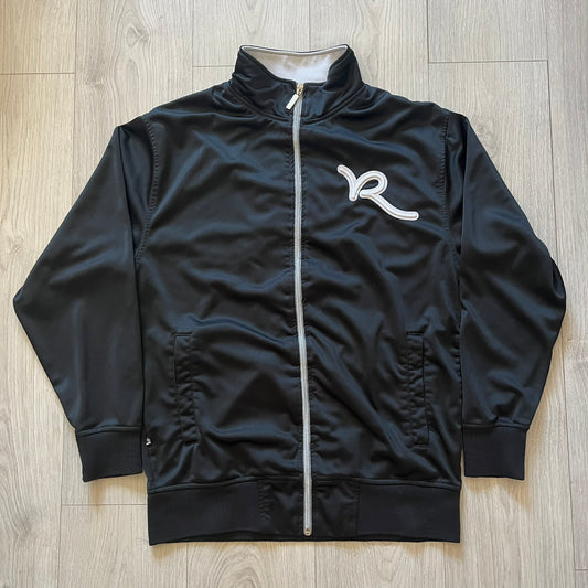 Rocawear black track jacket in size large, designed with a small embroidered logo on the chest and a large white logo across the back. A silver emblem near the wrist and a detailed silver zipper add distinctive touches. A sleek early 2000s streetwear piece.