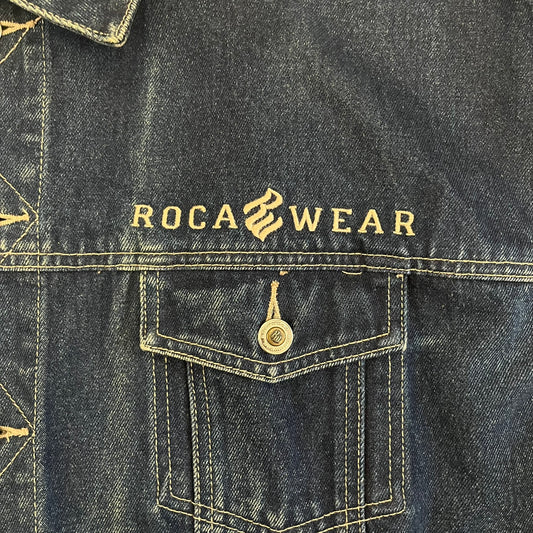 Rocawear blue denim jacket from the early 2000s, made from 100% cotton. Features an embroidered Rocawear logo on the right chest above the pocket, two buttoned chest pockets and standard side pockets. Finished with branded Rocawear metal buttons, a leather tab with metal R logo at the lower back, and “Rocawear All American Denim Industries” embroidery placed under the collar on the back. Classic structured denim fit with durable construction.
