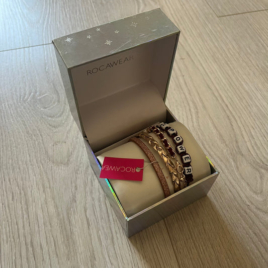 Set of four Rocawear bracelets, each with distinct design elements. Highlights include a “Girl Power” cube bracelet, a burgundy string with gold chain and pearl/zirconia charms, and two simple rose gold bands. Comes presented in a holographic silver box, ideal for collectors or as a stylish accessory set.