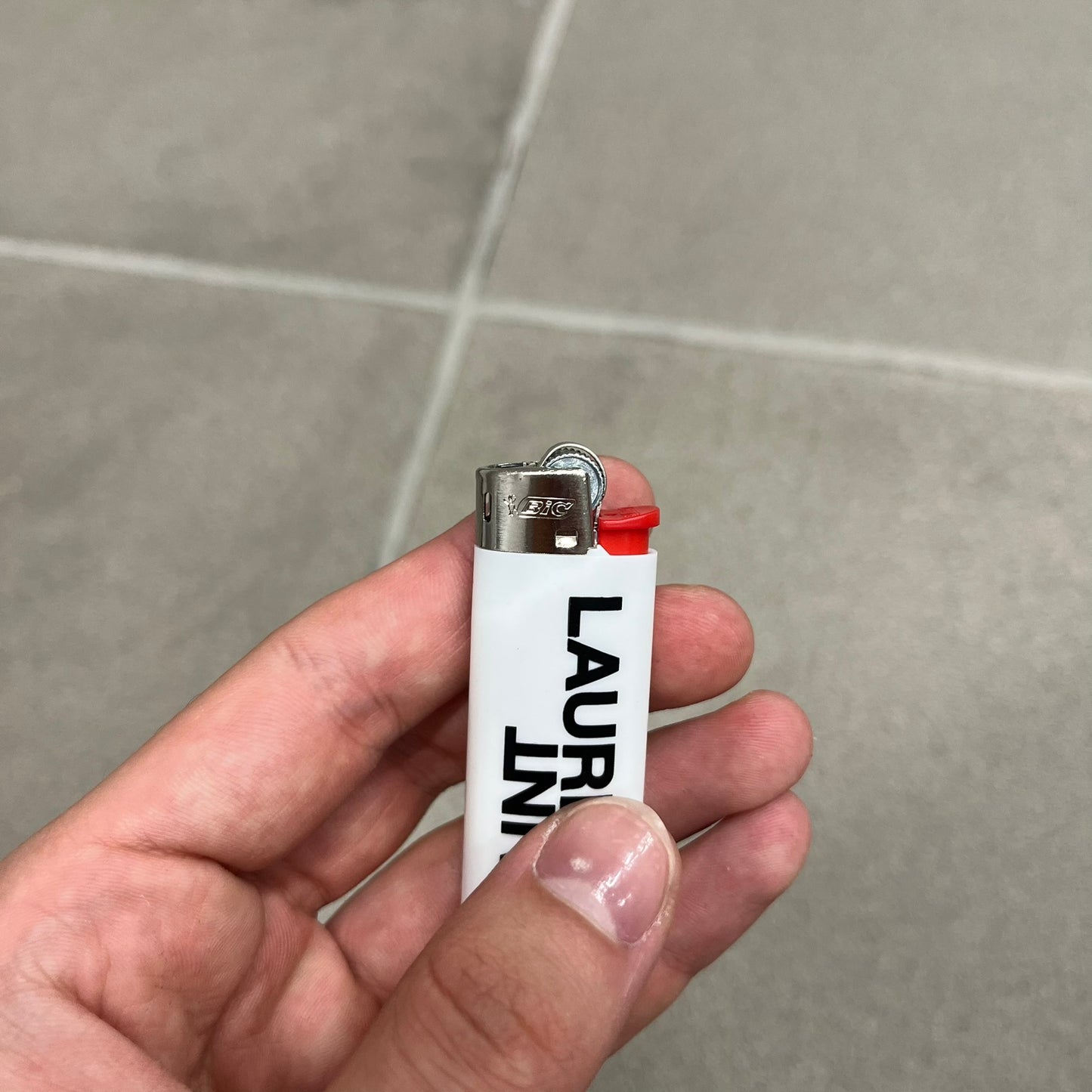 Saint Laurent White Logo Lighter is a white BIC gas lighter featuring black Saint Laurent branding, with “Saint” printed above and “Laurent” mirrored underneath. It is an in-store-only item made in Spain.