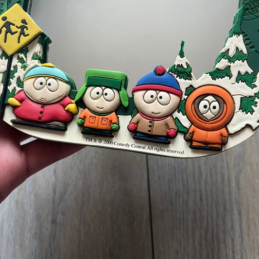 Small tabletop mirror with a molded rubber frame featuring all-over cutout logos for South Park and Comedy Central, along with leaf and tree motifs. The top is finished with a raised 3D South Park logo, while the bottom features 3D rubber figures of Cartman, Kenny, Kyle, and Stan standing at their bus stop. Marked with a 2000 Comedy Central copyright at the bottom, with a black backing and integrated stand.