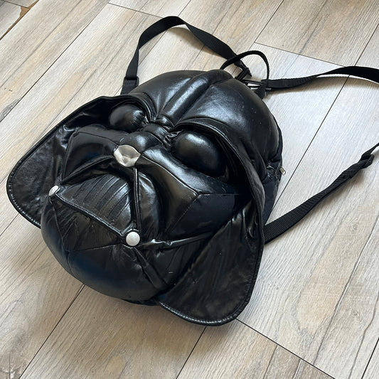 Star Wars 1990s Darth Vader Head Backpack is a vintage backpack shaped like Darth Vader’s helmet. It is finished in shiny black with molded plastic details recreating the character’s mask and helmet. The bag has one main compartment and includes a red Star Wars licensed tag from the 1990s.