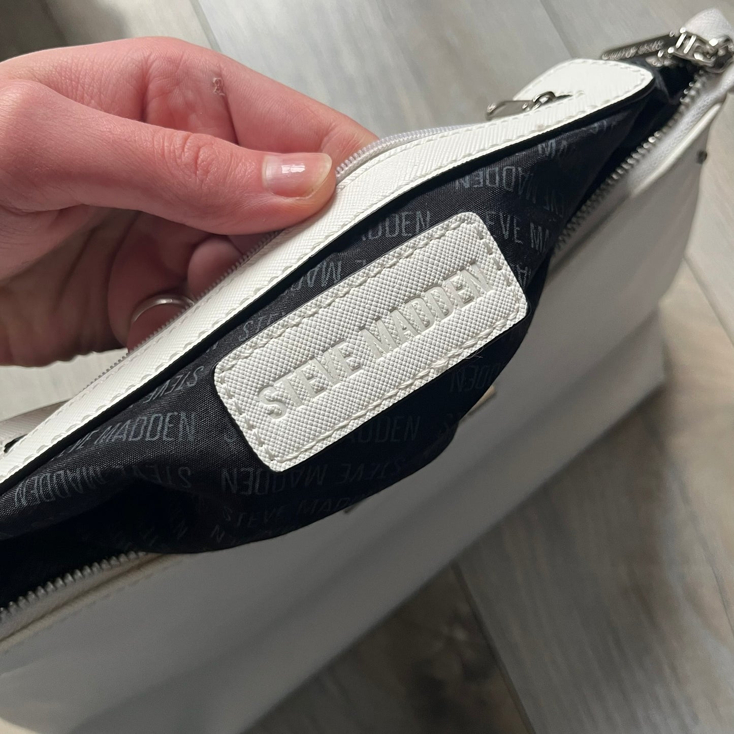 Steve Madden Removable Clutch White Handbag is a white leather tote featuring silver-tone hardware, including a front plaque and decorative buttons. The bag has two top handles and includes a removable inner clutch with silver Steve Madden branding. Designed for versatility, the clutch can be used separately or inside the main handbag.