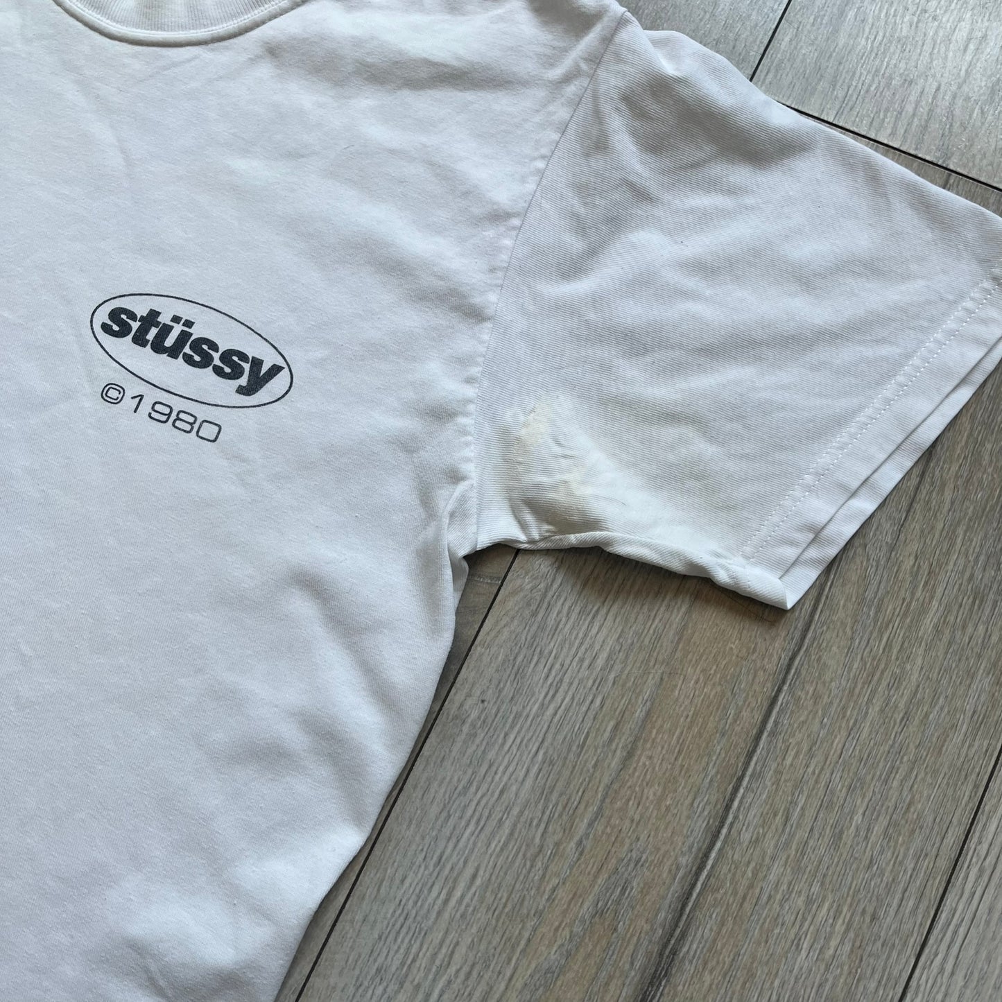 Stussy Soul 1980 White Tee is a white short-sleeve T-shirt released as part of a spring drop, made from soft cotton with a relaxed fit and ribbed collar. The front features a small Stussy logo print on the chest, while the back includes black Stussy text, a large multicolor central graphic, and “Copyright 1980” text at the bottom.