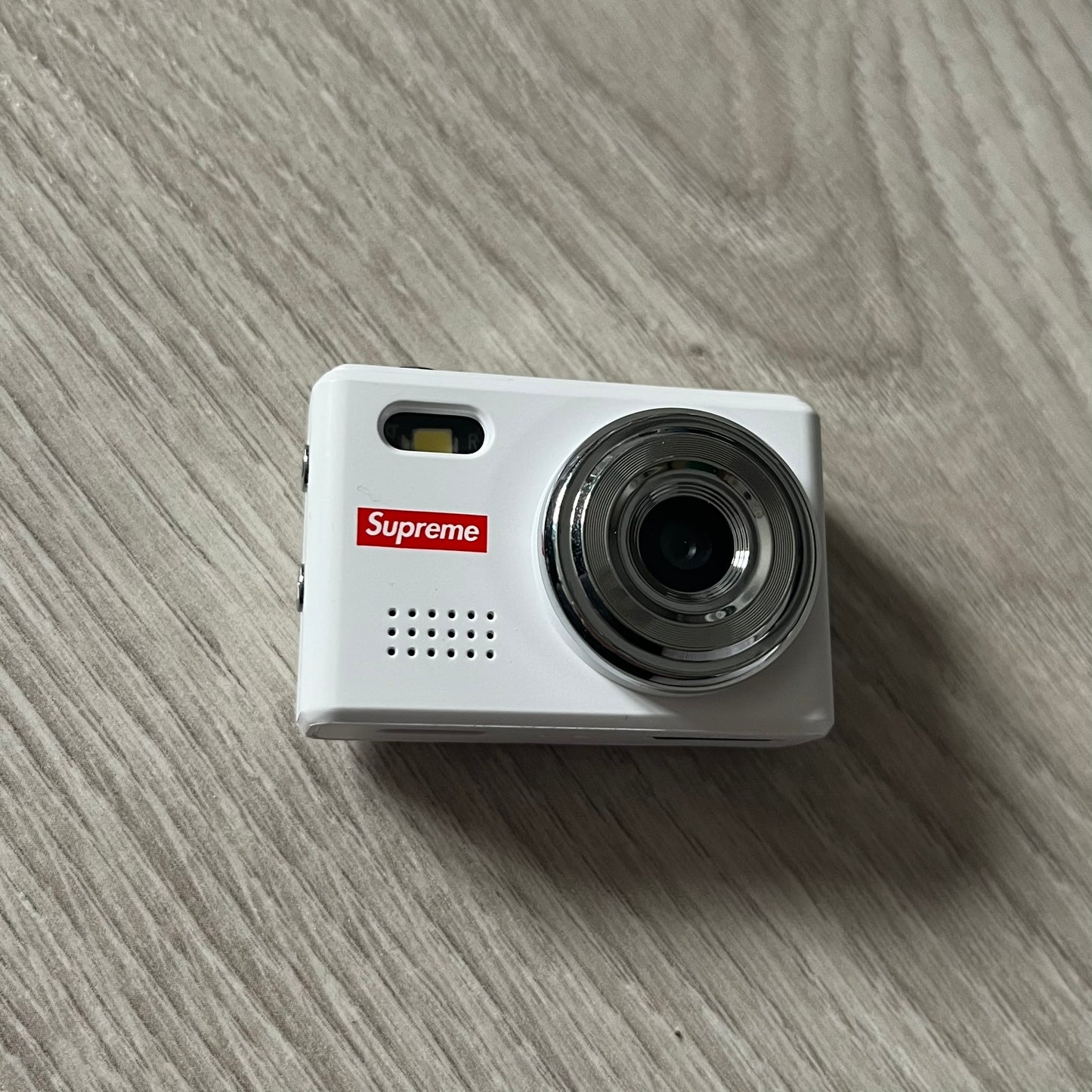 Supreme Digital Camera Keychain released for FW25 in white with a printed Supreme box logo on the front. This ultra-compact digital camera measures 1.92" x 1.14" x 1.1" and includes a 32GB TF card, USB-C cable, and built-in 1080p HD recording. When powered on, the screen displays the Supreme logo, with selectable interface colors in colors like: pink, green, or blue.