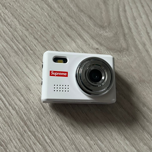 Supreme Digital Camera Keychain released for FW25 in white with a printed Supreme box logo on the front. This ultra-compact digital camera measures 1.92" x 1.14" x 1.1" and includes a 32GB TF card, USB-C cable, and built-in 1080p HD recording. When powered on, the screen displays the Supreme logo, with selectable interface colors in colors like: pink, green, or blue.