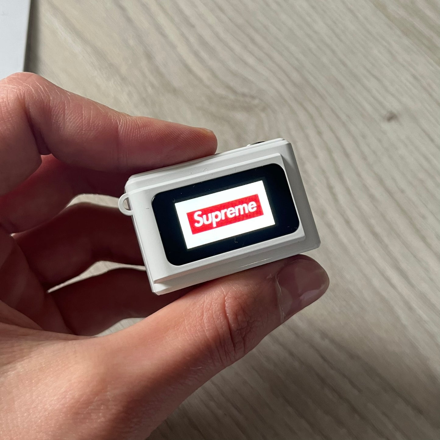 Supreme Digital Camera Keychain released for FW25 in white with a printed Supreme box logo on the front. This ultra-compact digital camera measures 1.92" x 1.14" x 1.1" and includes a 32GB TF card, USB-C cable, and built-in 1080p HD recording. When powered on, the screen displays the Supreme logo, with selectable interface colors in colors like: pink, green, or blue.