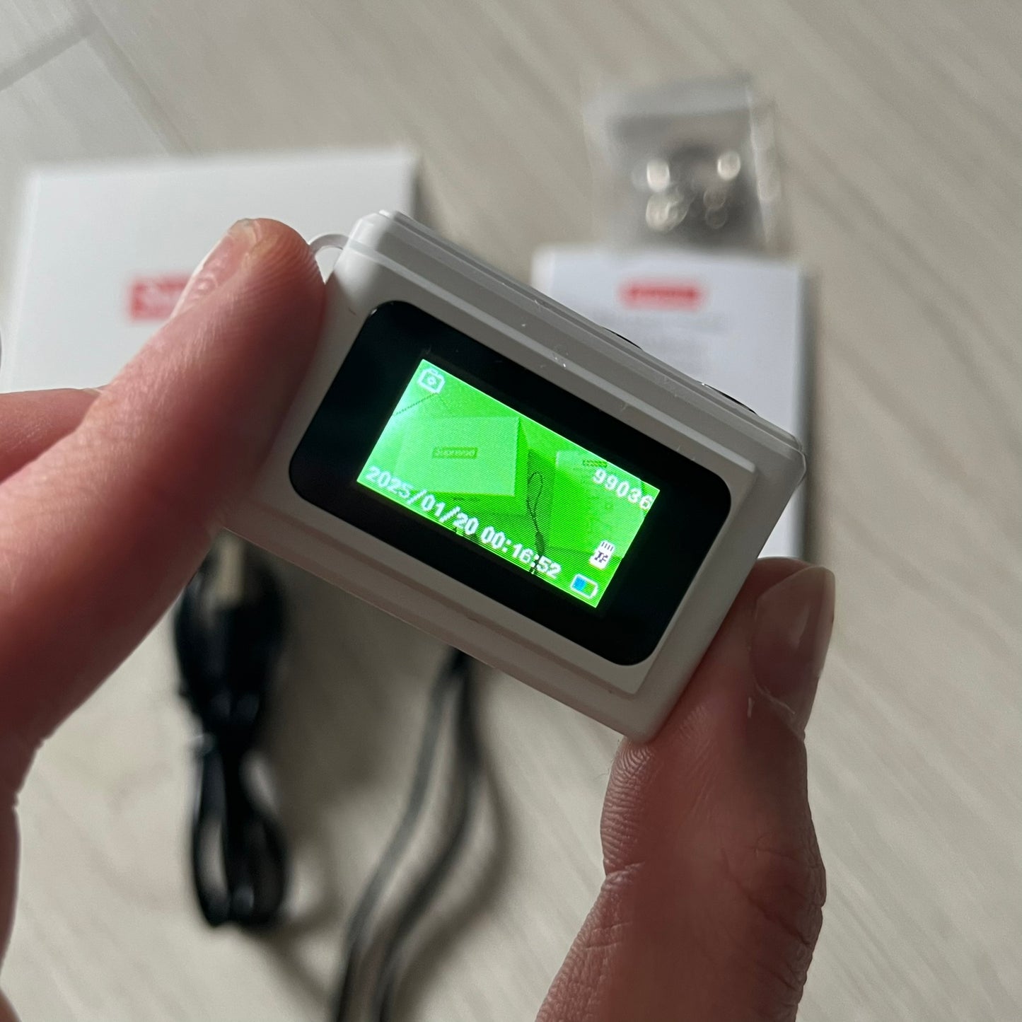 Supreme Digital Camera Keychain released for FW25 in white with a printed Supreme box logo on the front. This ultra-compact digital camera measures 1.92" x 1.14" x 1.1" and includes a 32GB TF card, USB-C cable, and built-in 1080p HD recording. When powered on, the screen displays the Supreme logo, with selectable interface colors in colors like: pink, green, or blue.