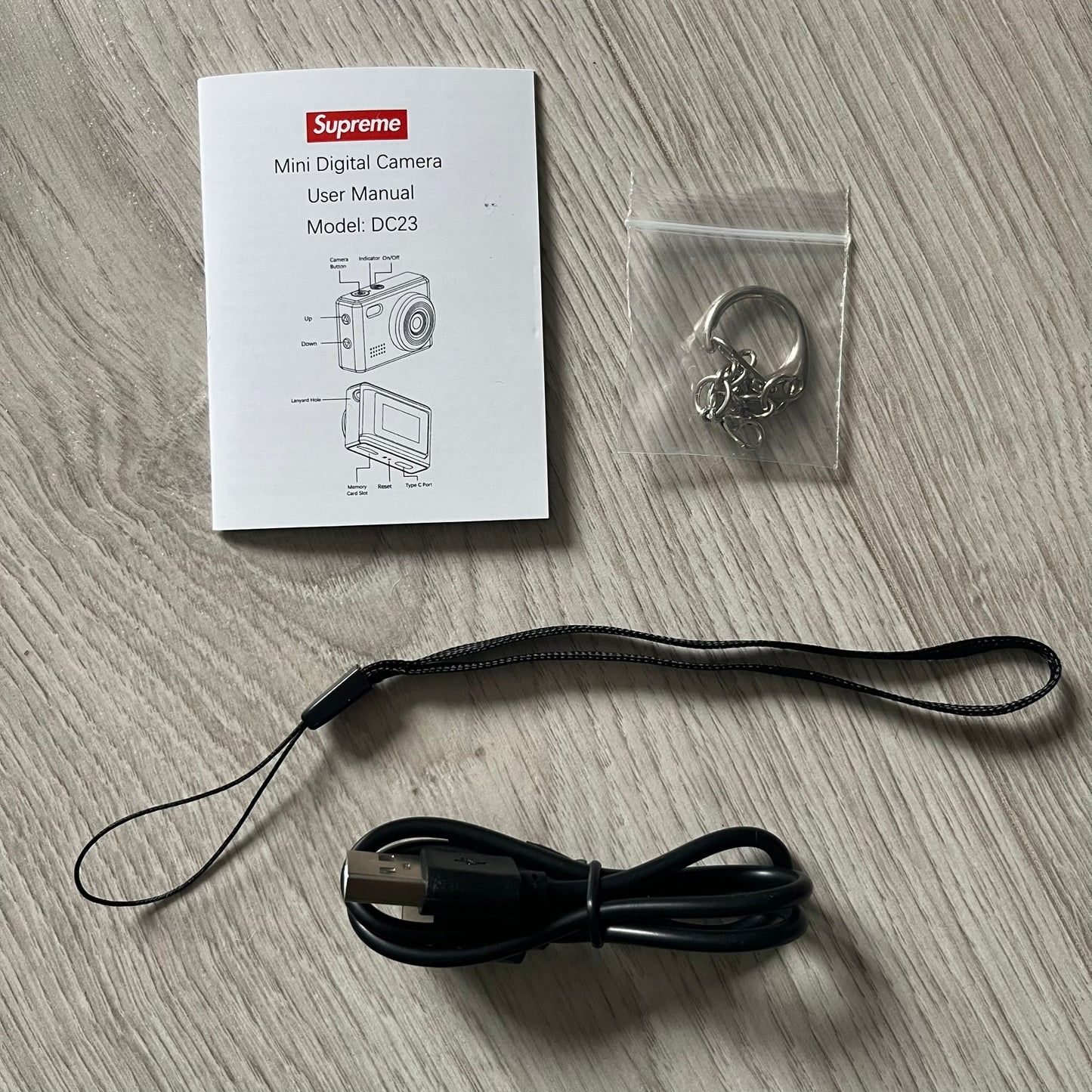 Supreme Digital Camera Keychain released for FW25 in white with a printed Supreme box logo on the front. This ultra-compact digital camera measures 1.92" x 1.14" x 1.1" and includes a 32GB TF card, USB-C cable, and built-in 1080p HD recording. When powered on, the screen displays the Supreme logo, with selectable interface colors in colors like: pink, green, or blue.