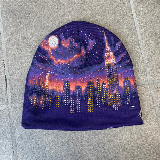 Supreme FW26 Cityscape Art Dealer Purple Beanie is a purple acrylic beanie with original artwork by Art Dealer, featuring a orange and light purple night city graphic with a moon and small stars across the sky on both sides. An old English-style Supreme logo sits on the front, and rhinestone appliqués in different sizes decorate the front and back. The print is stretchy, so any cracking effect that appears is just the print stretching with wear.