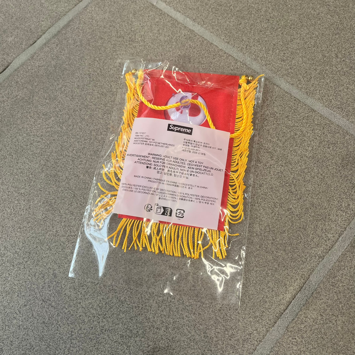 Supreme FW26 Fringed Banner is a small red banner trimmed with yellow fringe along the sides and bottom, with a yellow string at the top for hanging via suction mount. The design features a folded red box logo split across both sides of the banner, and it was distributed as a first-week free gift for FW26.