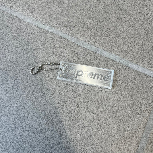 Supreme FW26 Reflective Clear Keychain is a clear plastic keychain on a steel ball chain, with a Supreme logo on the front. The back is made of small reflective panels.