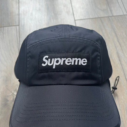 Supreme FW25 camp cap constructed from water-resistant Cordura® Advanced Fabrics nylon with a nylon and polyester shell. Lined with Polartec® 200 fleece for insulation, featuring packable earflaps secured with an interior elastic shockcord. Details include a webbing strap closure, 3M reflective piping, embroidered logos on the sides, and a classic Supreme box logo at the front. Finished in black.