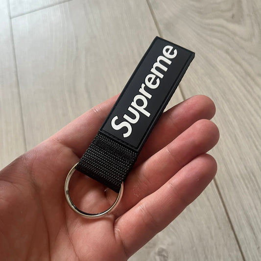 Supreme SS26 Webbing Keychain is a black keychain from the Spring/Summer 2026 collection. It features a silver-tone 1-inch keyring and a black rubber Supreme box logo patch attached to the front. The reverse side is plain with no additional graphics.