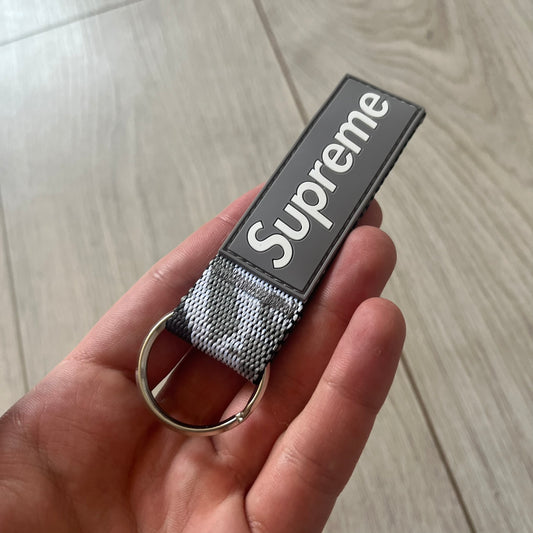 Supreme SS26 Webbing Keychain is a gray keychain from the Spring/Summer 2026 collection. It features a silver-tone 1-inch keyring and a gray rubber Supreme box logo patch attached to the front. The reverse side is a gray, white and black "snow camo".