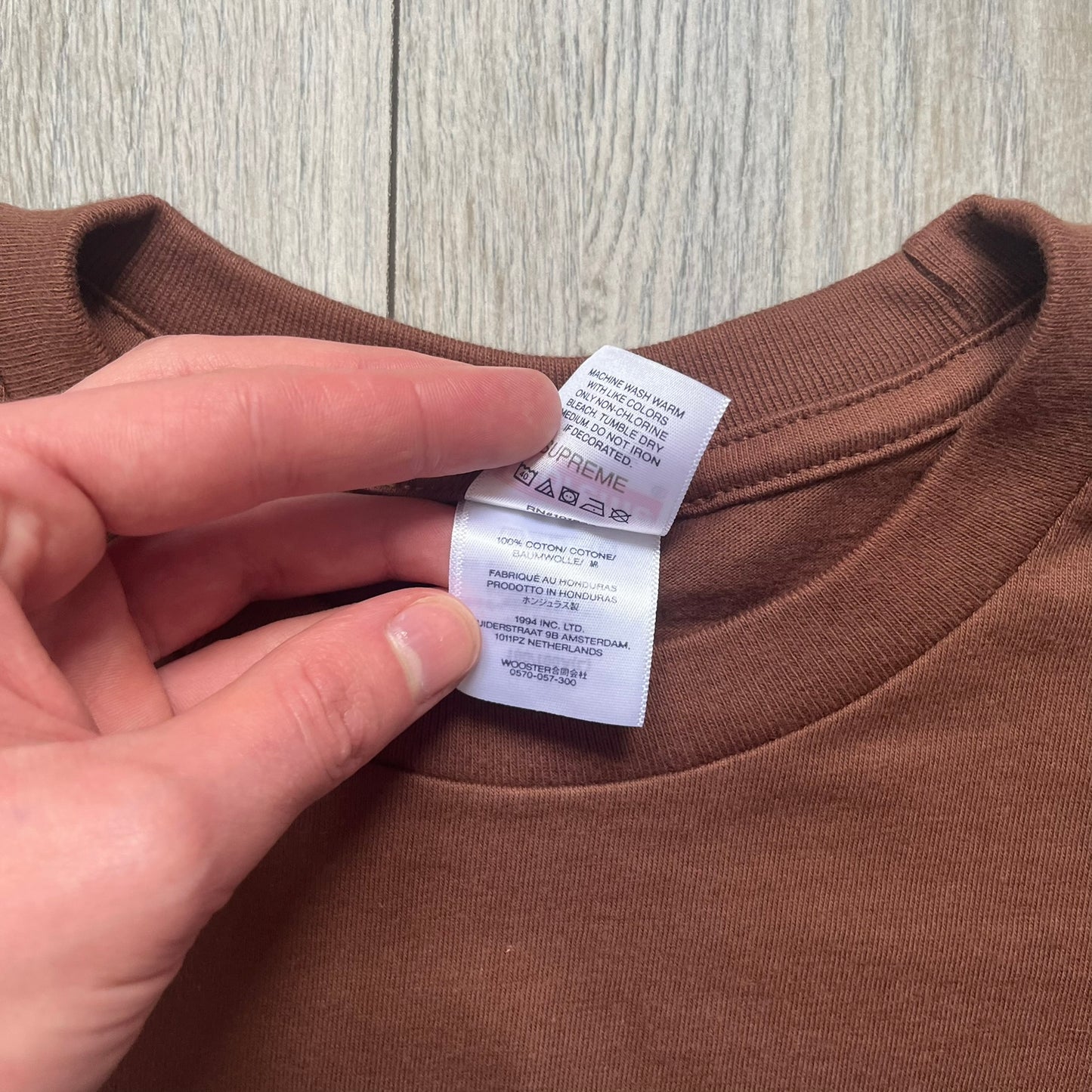 Supreme x Spider-Man Black Tee from the SS26 season is a brown 100% cotton T-shirt featuring a large box graphic on the front. The print shows a black and white city photo with the Supreme store, while Spider-Man in his red and blue costume swings across the scene in a cartoon-style illustration. The shirt includes a Supreme x Marvel collaboration neck tag.