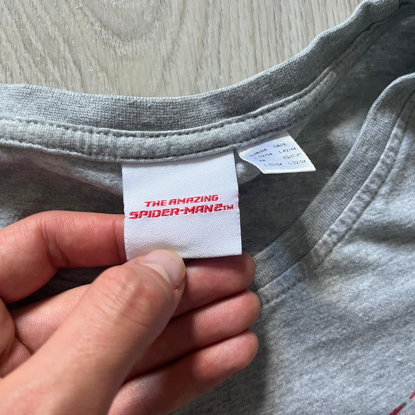 Gray 2014 Marvel tee featuring The Amazing Spider-Man 2 design with a “ripped page” effect showcasing Spider-Man’s face in the center. Text logo sits beneath the graphic for a bold cinematic touch. A collectible piece from the official movie merchandise line.