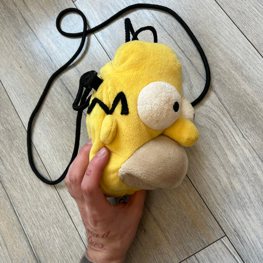The Simpsons 1990s Homer Simpson Head Shoulder bag is shaped like a 3D Homer Simpson head. The design is yellow with soft sculpted facial features including eyes and nose, a molded beige mouth, and protruding ears. The hair detail wraps around the back, with the top strands functioning as a hanger. It includes a black string and a single main compartment.