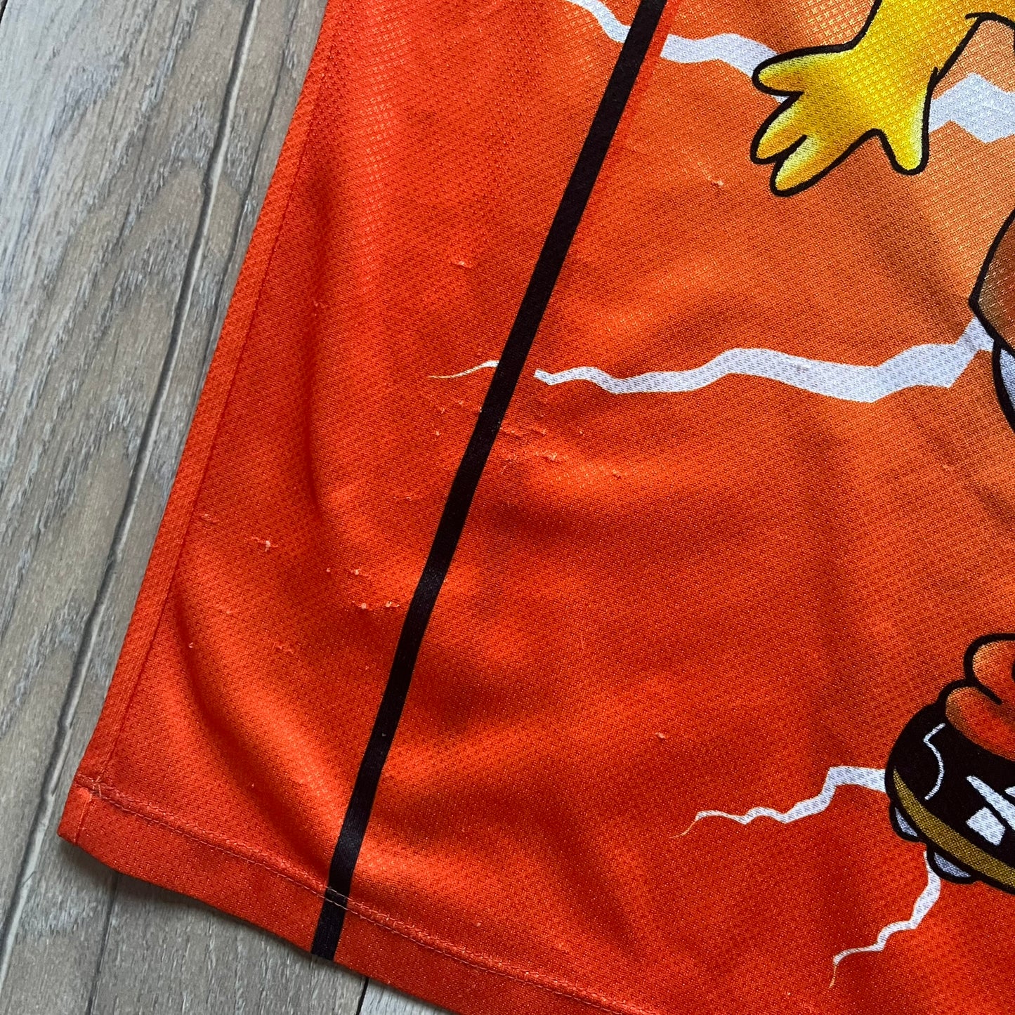 Officially licensed 1999 The Simpsons World Cup Netherlands Tee. Orange lightweight polyester tee with an all-over graphic design. The front features Bart Simpson kicking a football, orange “Bart” text, white lightning graphics, and a Netherlands emblem with Bart illustrated beneath it. Black flame details on the sleeve, along with a black ribbed collar with orange lining. Matt Groening / The Simpsons ©1999 licensing is printed below the main graphic. The back repeats the design in a flipped layout, showing