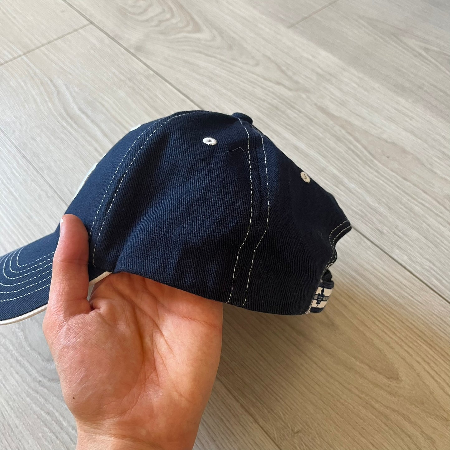 Hand holding a navy blue cap with white stitching on a light wooden floor.