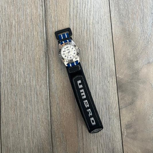 Umbro U528 The Double Diamond Analog Watch features a white dial with navy Umbro logo and numerals, silver and navy hands, and a silver case. The watch comes with a black Velcro strap accented with large white Umbro text and a small blue-and-white detail. It combines sporty design with a straightforward analog display.
