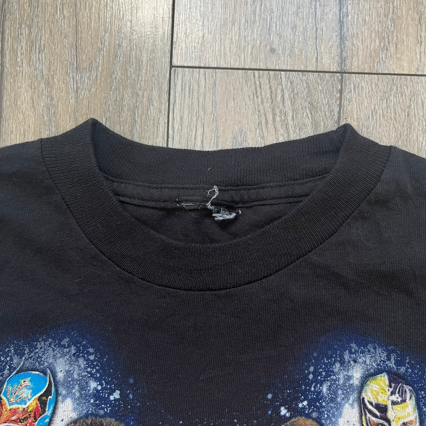 WWE 2011 All Over Print Promo Tee is a black short-sleeve T-shirt featuring a large front graphic of multiple WWE wrestlers, including Rey Mysterio, John Cena, and Randy Orton. A small WWE logo is printed beneath the main graphic. The shirt has no neck tag and was released as official promotional merchandise in 2011.