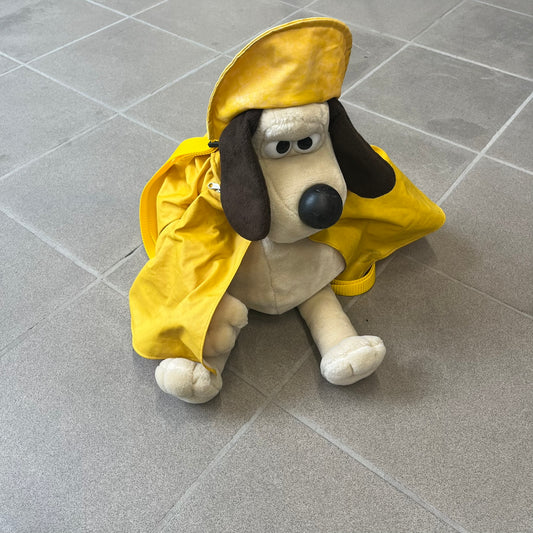 The Wallace and Gromit Gromit Plush Backpack is a 1990s licensed item produced by Aardman Animations. It features Gromit dressed in his bright yellow PVC raincoat and matching hat, as seen in The Wrong Trousers. The backpack is made from soft plush fabric with plastic eyes, a hard black nose, and a genuine stamp confirming official production. The bag opens from the side and includes adjustable shoulder straps for wearability.
