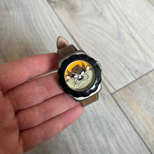 Warner Bros Tasmanian Devil 1997 Analog Watch is a stainless steel watch with a black-silver case and a fresh brown leather strap. The dial features a graphic of Taz smiling in front of a orange background. The caseback includes Warner Bros 1997 licensing
