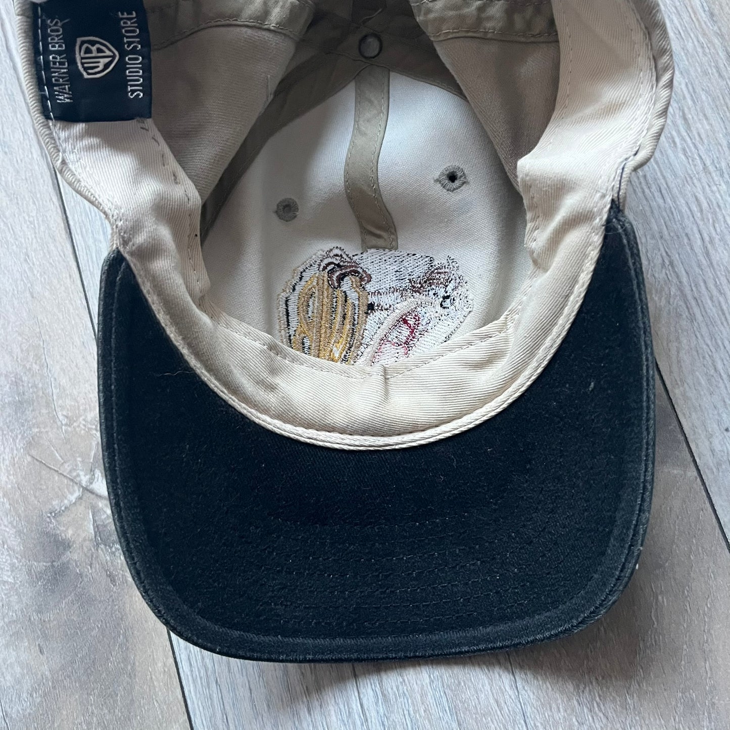Warner Bros Taz 1998 Baseball Cap is a two-tone beige and black cap featuring an embroidered Warner Bros logo on the front, positioned next to a detailed Taz (Tasmanian Devil) character graphic. Inside, the cap retains its original 1998 Warner Bros tag. The adjustable back strap has been reattached and stitched.