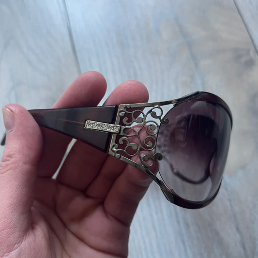 The Yves Saint Laurent 2007 Swirls 6412/S Sunglasses are an oversized model from 2007 with Category 3 sun protection. They feature oversized gradient grey lenses and burgundy (bordeaux) plastic frames, with silver-tone metal swirl details connecting the lenses to the temples. The temples are finished with a metal Yves Saint Laurent logo integrated into the swirl hardware. Made in Italy, with all original markings located on the inside. They come with a white leather YSL case.