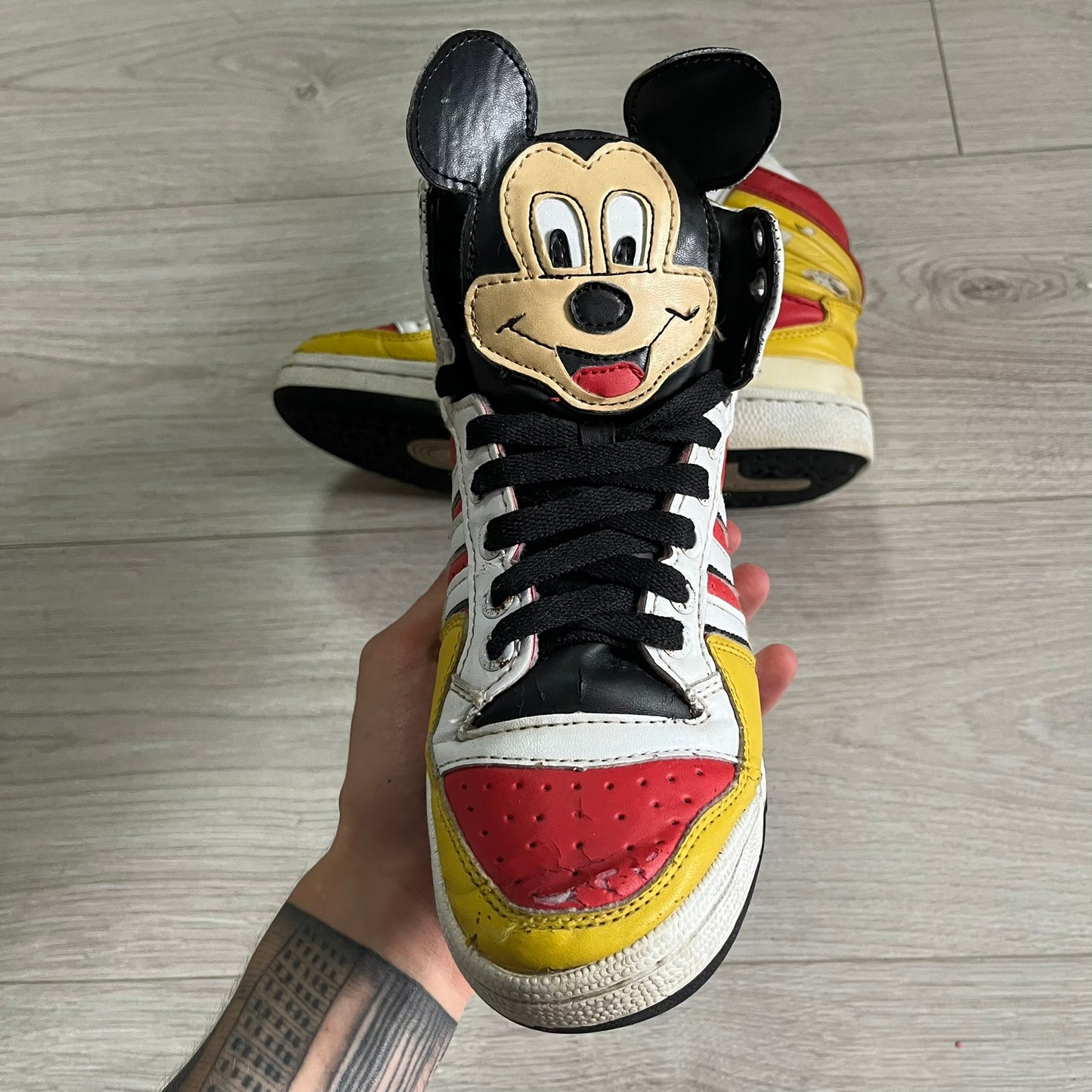 Sneakers with a cartoon face design held by a hand on a wooden floor.