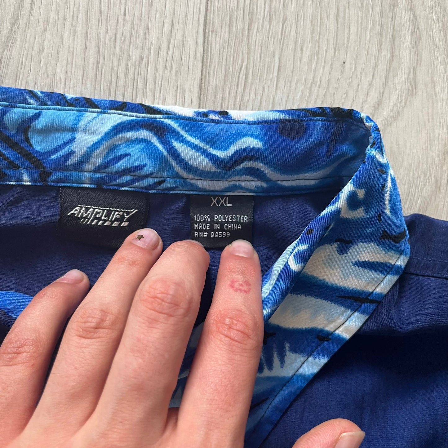 Hand holding a blue patterned shirt with an 'Amplify' label on a wooden surface