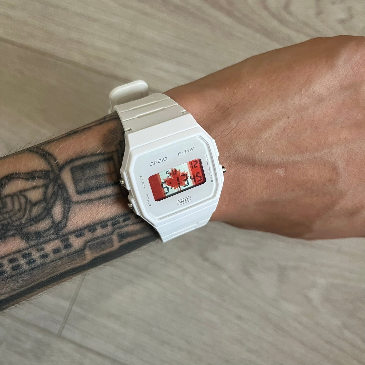 White Casio watch on a wrist with a red face, worn by a person with tattoos.