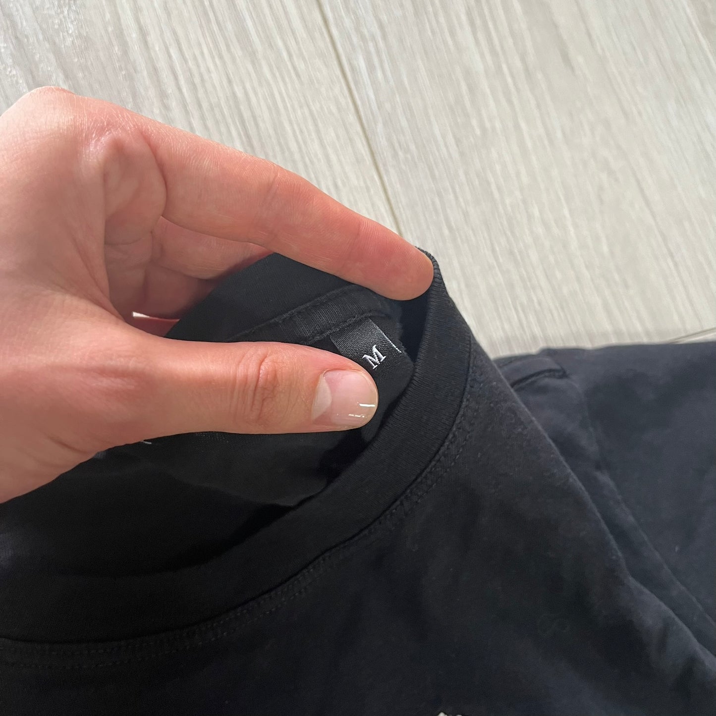 Hand pointing to a size label on a black garment against a light wooden floor background