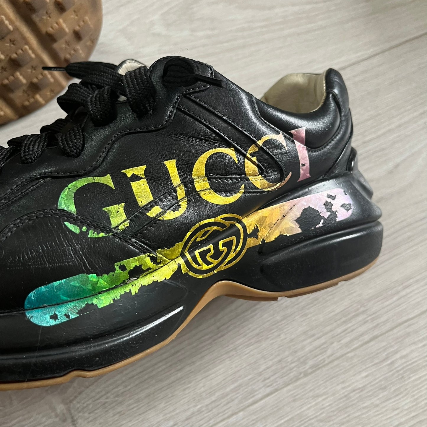 Black Gucci sneaker with colorful design on a light wooden floor.