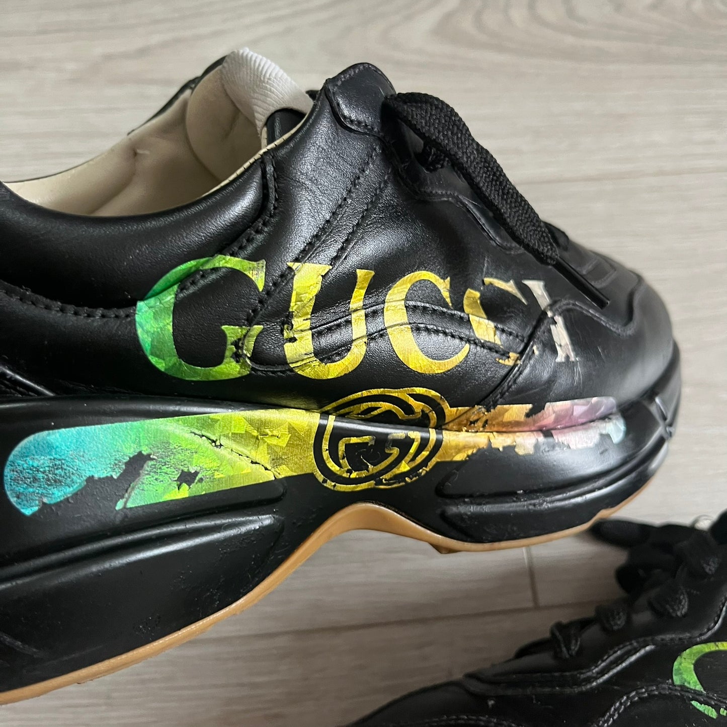 Black sneaker with colorful 'Gucci' logo on a light wooden floor.