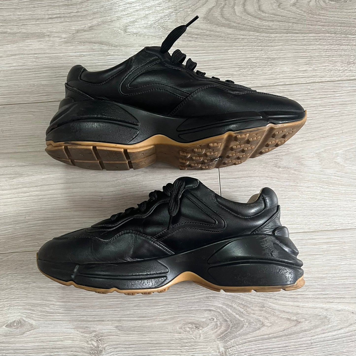 Pair of black sneakers with brown soles on a light wooden floor.