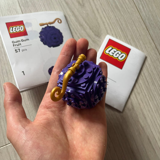 Hand holding a purple LEGO model of a gum ball with a gold stick, with LEGO packaging in the background.