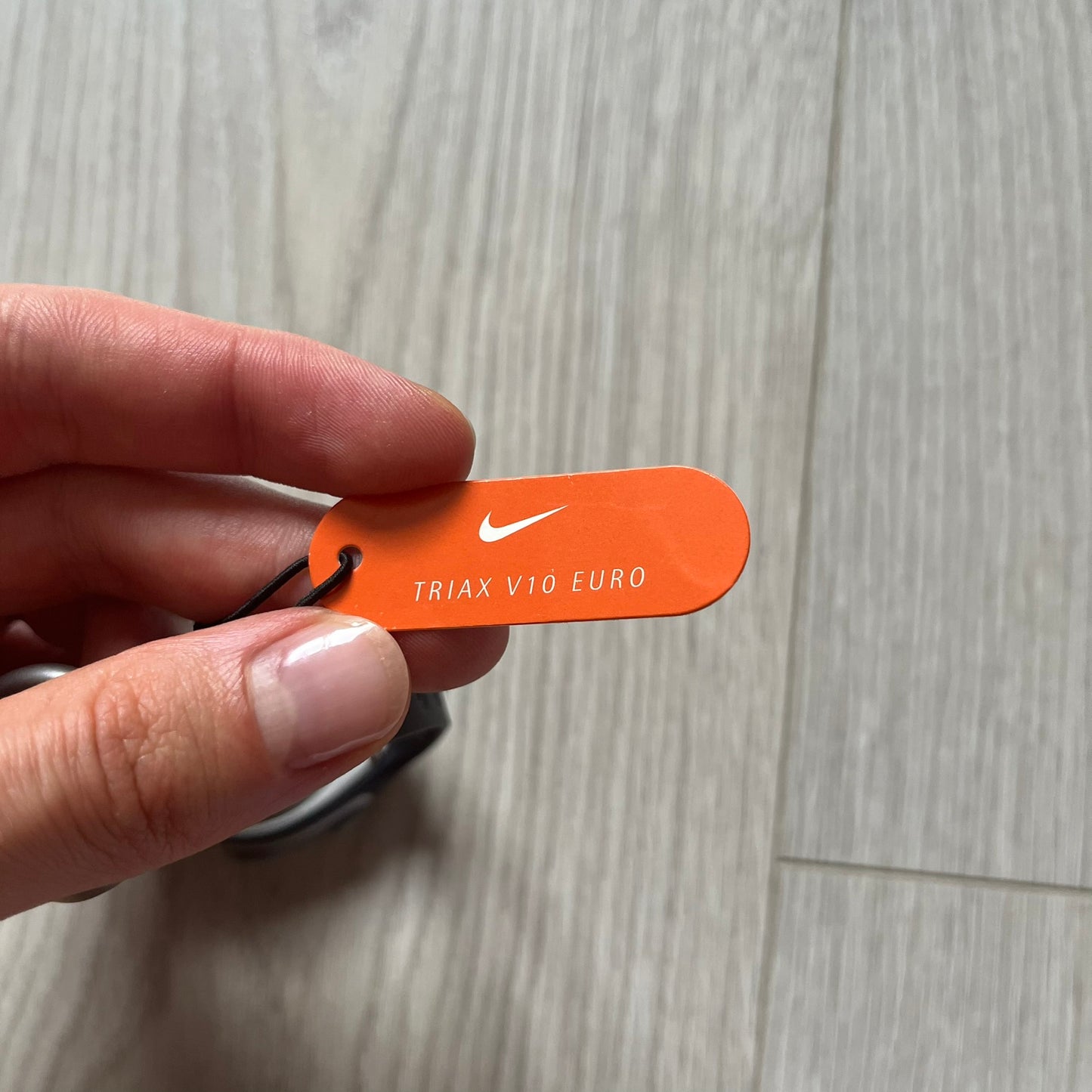 Hand holding an orange Nike tag with 'TRIAX V10 EURO' text on a light wooden floor.